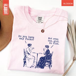 May include: A light pink t-shirt with the text "Do you have any fucks?" and "Not even one, girl. Go fish." printed in navy blue. The shirt features a graphic of two women. A gold necklace is draped on the shirt.