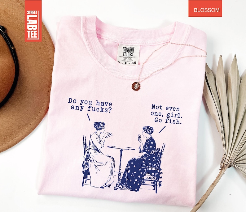 May include: A light pink t-shirt with the text "Do you have any fucks?" and "Not even one, girl. Go fish." printed in navy blue. The shirt features a graphic of two women. A gold necklace is draped on the shirt.