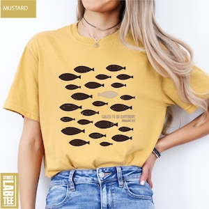 Puede incluir: Camiseta de color mostaza con un gr&aacute;fico de peces, uno de los cuales es de un color diferente. El texto "CALLED TO BE DIFFERENT" y "ROMANS 12:2" est&aacute; impreso en la camiseta. La camiseta se combina con unos vaqueros azules.