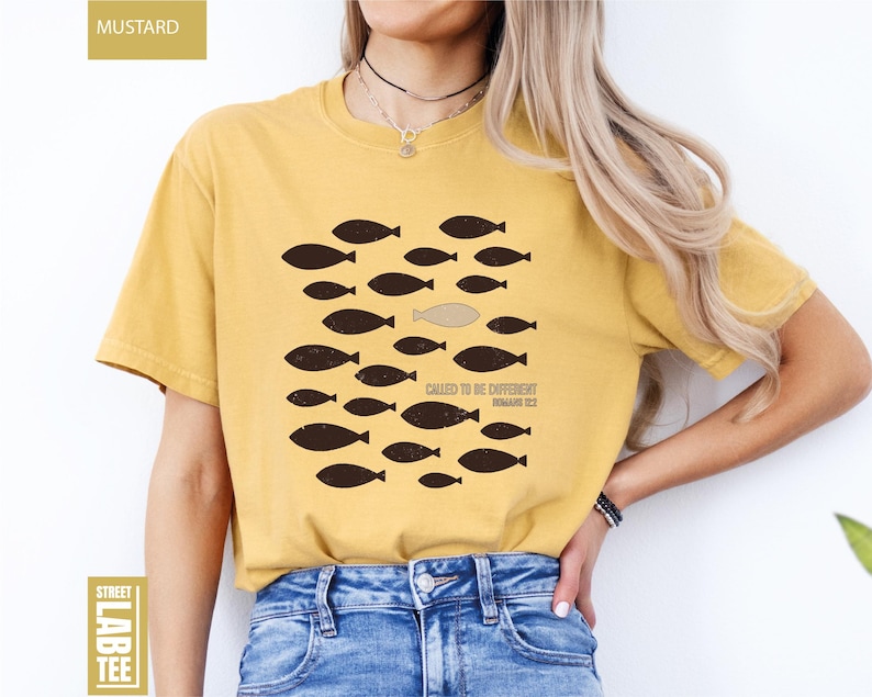 Puede incluir: Camiseta de color mostaza con un gr&aacute;fico de peces, uno de los cuales es de un color diferente. El texto "CALLED TO BE DIFFERENT" y "ROMANS 12:2" est&aacute; impreso en la camiseta. La camiseta se combina con unos vaqueros azules.