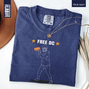 Op de afbeelding: Een marineblauw t-shirt met de tekst "FREE DC" en een afbeelding van een American footballspeler die een bal gooit. Het shirt heeft een ronde hals en korte mouwen. Een gouden ketting is over het shirt gedrapeerd.