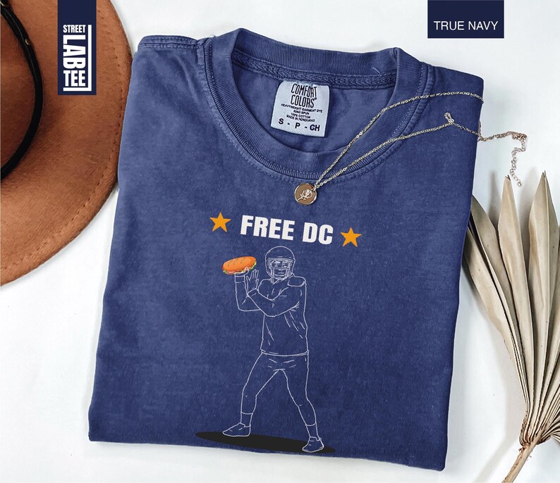 Op de afbeelding: Een marineblauw t-shirt met de tekst "FREE DC" en een afbeelding van een American footballspeler die een bal gooit. Het shirt heeft een ronde hals en korte mouwen. Een gouden ketting is over het shirt gedrapeerd.