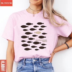 Puede incluir: Camiseta rosa claro con un dise&ntilde;o de peces, uno de ellos de diferente color. El texto "CALLED TO BE DIFFERENT ROMANS 12:2" est&aacute; impreso debajo del dise&ntilde;o. La camiseta tiene mangas cortas.