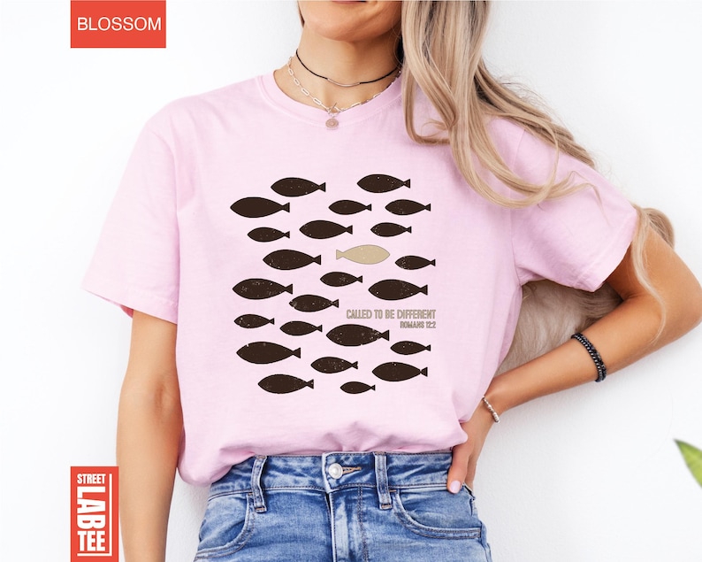 Puede incluir: Camiseta rosa claro con un dise&ntilde;o de peces, uno de ellos de diferente color. El texto "CALLED TO BE DIFFERENT ROMANS 12:2" est&aacute; impreso debajo del dise&ntilde;o. La camiseta tiene mangas cortas.
