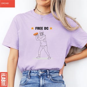 Op de afbeelding: Lichtpaars T-shirt met de tekst "FREE DC" en een afbeelding van een American footballspeler die een bal gooit. Het shirt heeft een ronde hals en korte mouwen.
