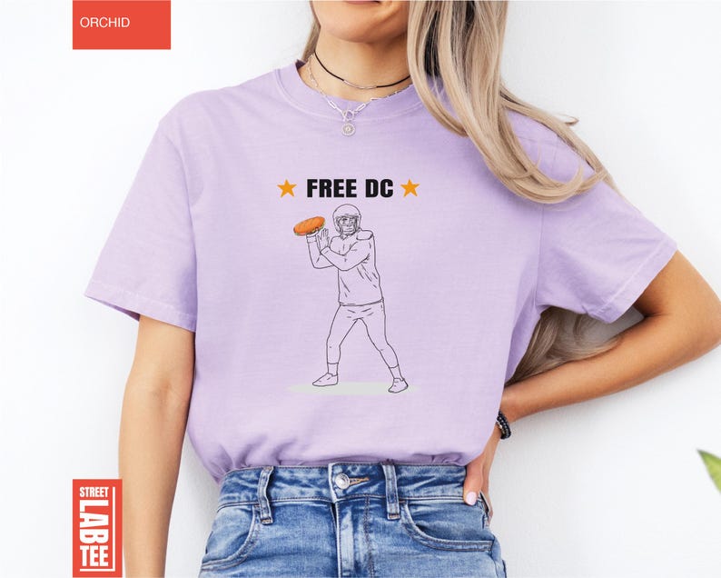 Op de afbeelding: Lichtpaars T-shirt met de tekst "FREE DC" en een afbeelding van een American footballspeler die een bal gooit. Het shirt heeft een ronde hals en korte mouwen.
