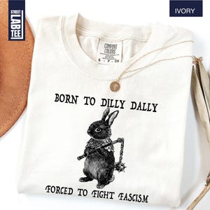 May include: Ivory-colored t-shirt with the text "BORN TO DILLY DALLY" above a black and white illustration of a rabbit holding a mace. Below the rabbit is the text "FORCED TO FIGHT FASCISM". The shirt has a crew neck and short sleeves.