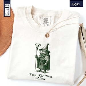 May include: Ivory-colored t-shirt featuring a green illustration of a frog dressed as a wizard, holding a staff. The text "Twas The 'Tism M'lord" is printed below the illustration. The shirt has a crew neck and short sleeves.