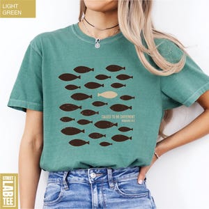 Puede incluir: Camiseta verde claro con un dise&ntilde;o de peces marrones y el texto "CALLED TO BE DIFFERENT" y "ROMANS 12:2". Un estilo casual y c&oacute;modo.