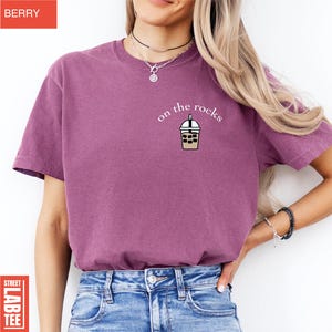 May include: Berry-colored t-shirt with the phrase "on the rocks" and a graphic of an iced coffee drink. The shirt is a short-sleeved crew neck style. The shirt is made of a soft material.
