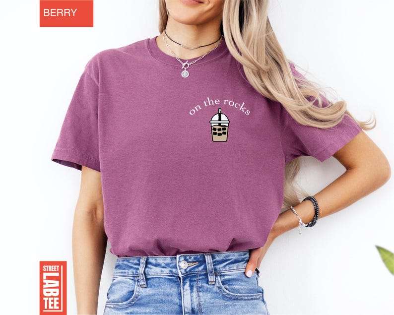 May include: Berry-colored t-shirt with the phrase "on the rocks" and a graphic of an iced coffee drink. The shirt is a short-sleeved crew neck style. The shirt is made of a soft material.