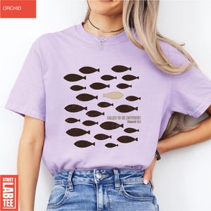 Puede incluir: Camiseta de color lila con un dise&ntilde;o de peces marrones, un pez beige y el texto "CALLED TO BE DIFFERENT" y "ROMANS 12:2". La camiseta es una Street Lab Tee. La palabra "ORCHID" est&aacute; impresa en un rect&aacute;ngulo rojo en la esquina superior izquierda.