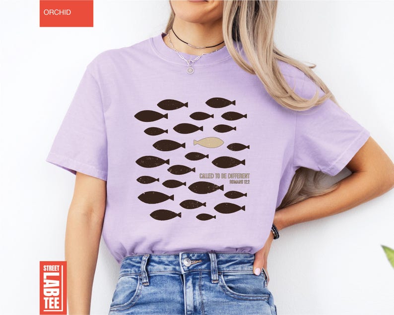 Puede incluir: Camiseta de color lila con un dise&ntilde;o de peces marrones, un pez beige y el texto "CALLED TO BE DIFFERENT" y "ROMANS 12:2". La camiseta es una Street Lab Tee. La palabra "ORCHID" est&aacute; impresa en un rect&aacute;ngulo rojo en la esquina superior izquierda.