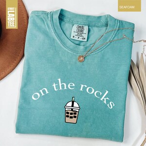 May include: Seafoam green t-shirt with the phrase "on the rocks" and an iced coffee graphic. The shirt is Comfort Colors brand. A gold necklace with a pendant rests on the shirt. The shirt is a seafoam green colour.
