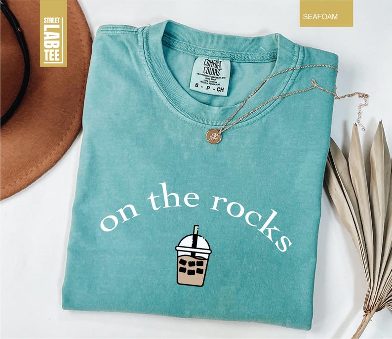 May include: Seafoam green t-shirt with the phrase "on the rocks" and an iced coffee graphic. The shirt is Comfort Colors brand. A gold necklace with a pendant rests on the shirt. The shirt is a seafoam green colour.