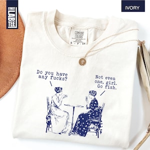 May include: Ivory-colored t-shirt with a blue graphic of two women and the text "Do you have any fucks?" and "Not even one, girl. Go fish." A gold necklace is draped on the shirt. The shirt has a Comfort Colors tag.