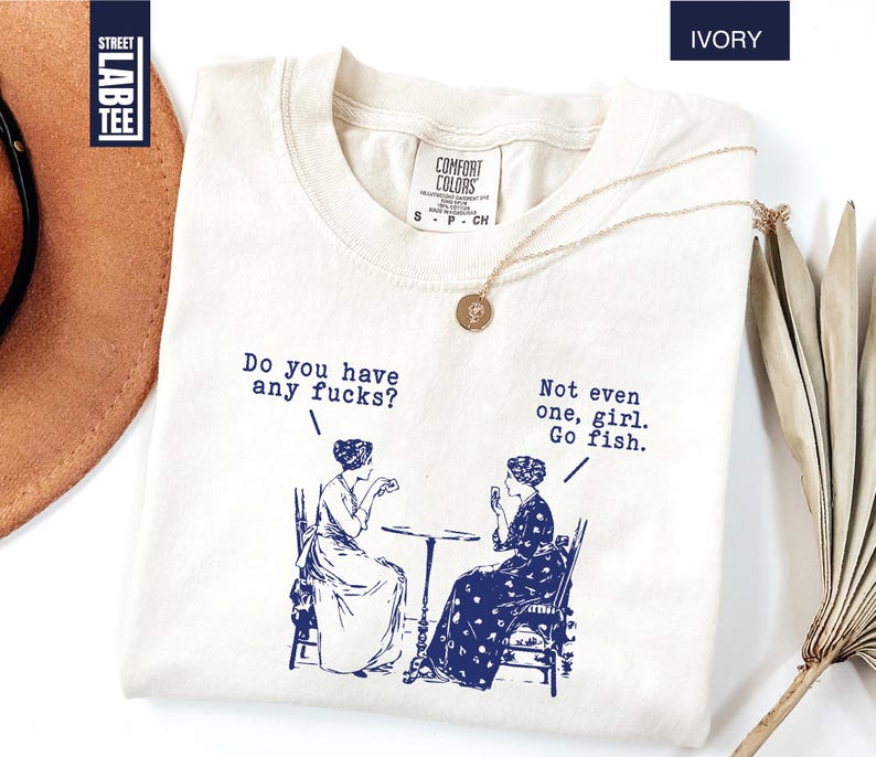May include: Ivory-colored t-shirt with a blue graphic of two women and the text "Do you have any fucks?" and "Not even one, girl. Go fish." A gold necklace is draped on the shirt. The shirt has a Comfort Colors tag.