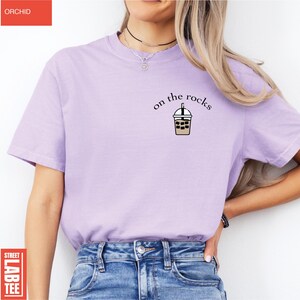 May include: A light purple t-shirt with the text "on the rocks" above a graphic of an iced beverage. The shirt is a solid color and has short sleeves. The word "ORCHID" is in a red rectangle in the upper left corner.