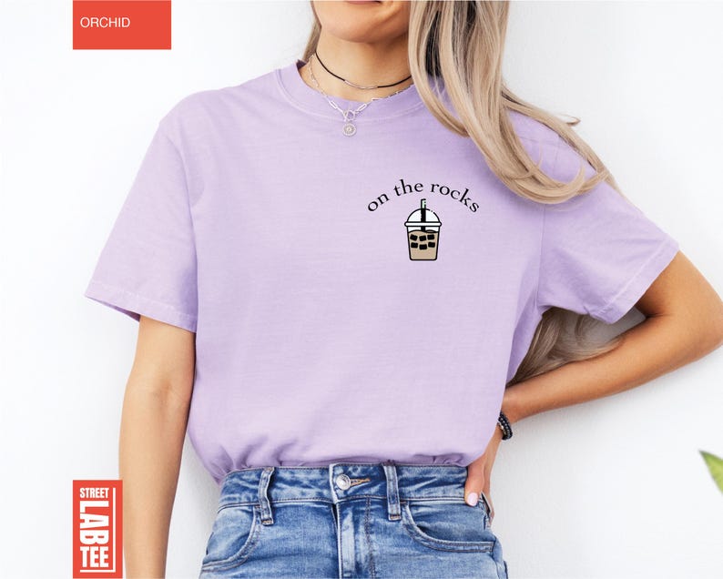 May include: A light purple t-shirt with the text "on the rocks" above a graphic of an iced beverage. The shirt is a solid color and has short sleeves. The word "ORCHID" is in a red rectangle in the upper left corner.