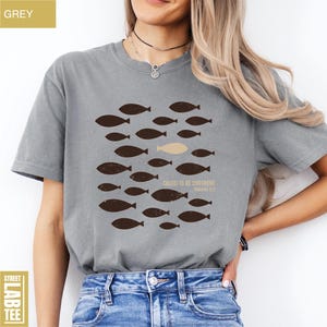 Puede incluir: Camiseta gris con un dise&ntilde;o de peces. El dise&ntilde;o presenta un grupo de peces marrones nadando juntos, con un pez beige que destaca. El texto "CALLED TO BE DIFFERENT" y "ROMANS 12:2" est&aacute; impreso debajo.