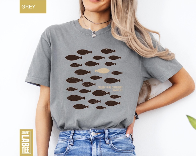 Puede incluir: Camiseta gris con un dise&ntilde;o de peces. El dise&ntilde;o presenta un grupo de peces marrones nadando juntos, con un pez beige que destaca. El texto "CALLED TO BE DIFFERENT" y "ROMANS 12:2" est&aacute; impreso debajo.