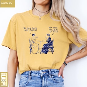 May include: Mustard-colored t-shirt with a humorous graphic in navy blue. The graphic depicts two women seated at a table with speech bubbles. One bubble reads, "Do you have any fucks?" and the other says, "Not even one, girl. Go fish."