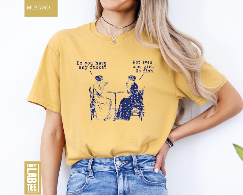 May include: Mustard-colored t-shirt with a humorous graphic in navy blue. The graphic depicts two women seated at a table with speech bubbles. One bubble reads, "Do you have any fucks?" and the other says, "Not even one, girl. Go fish."