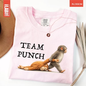 May include: A light pink t-shirt with the words "TEAM PUNCH" in black. The shirt features a cartoon illustration of two monkeys, one pulling the other. The shirt is made of Comfort Colors material.