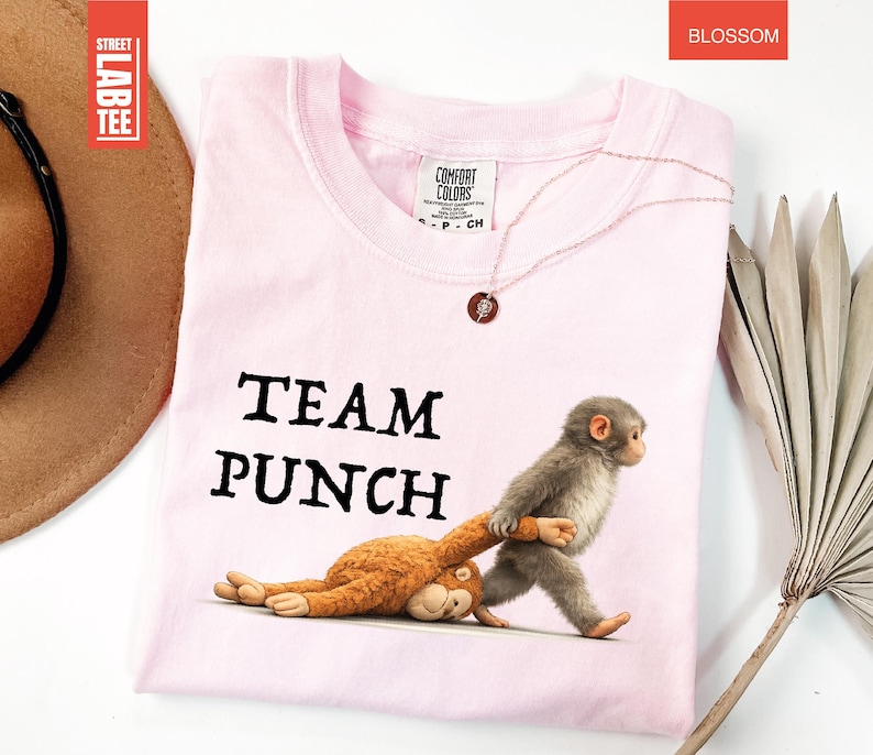 May include: A light pink t-shirt with the words "TEAM PUNCH" in black. The shirt features a cartoon illustration of two monkeys, one pulling the other. The shirt is made of Comfort Colors material.