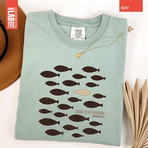 Puede incluir: Camiseta azul claro con un dise&ntilde;o de peces. El dise&ntilde;o presenta un grupo de peces marrones oscuros nadando juntos, con un pez beige nadando en la direcci&oacute;n opuesta. El texto "CHASED TO BE DIFFERENT" est&aacute; impreso debajo. Un collar dorado est&aacute; sobre la camiseta.