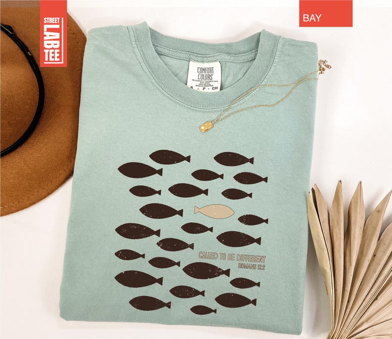 Puede incluir: Camiseta azul claro con un dise&ntilde;o de peces. El dise&ntilde;o presenta un grupo de peces marrones oscuros nadando juntos, con un pez beige nadando en la direcci&oacute;n opuesta. El texto "CHASED TO BE DIFFERENT" est&aacute; impreso debajo. Un collar dorado est&aacute; sobre la camiseta.