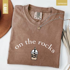 May include: Espresso-coloured t-shirt featuring the words "on the rocks" in white script and an iced drink graphic. The shirt has a crew neck and a small tag. A silver necklace with a pendant is on the shirt.