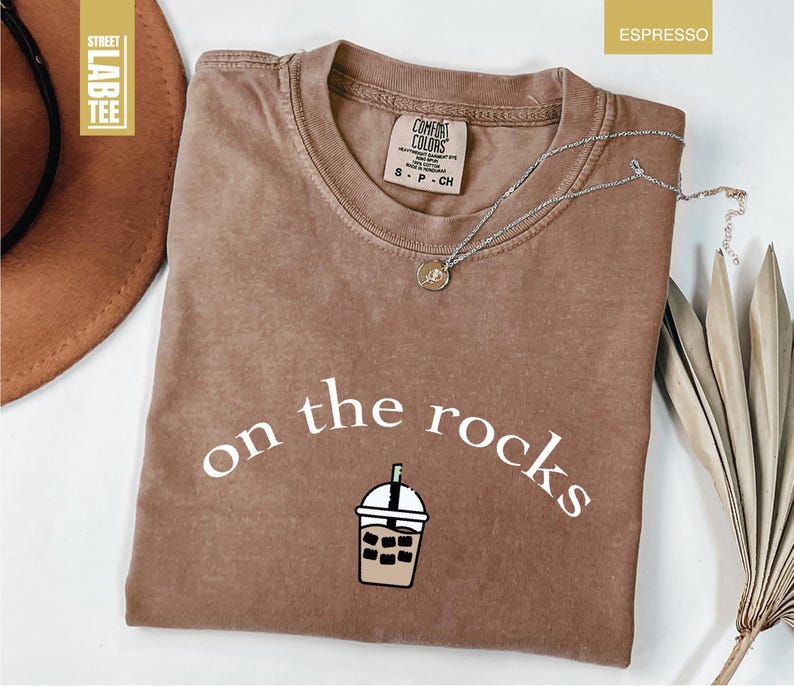 May include: Espresso-coloured t-shirt featuring the words "on the rocks" in white script and an iced drink graphic. The shirt has a crew neck and a small tag. A silver necklace with a pendant is on the shirt.