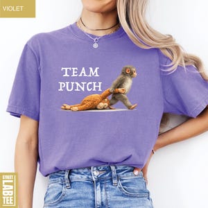 May include: A violet-colored t-shirt with the words "TEAM PUNCH" printed in white. The shirt features a graphic of a monkey pulling a stuffed animal. The shirt is made of a soft material.