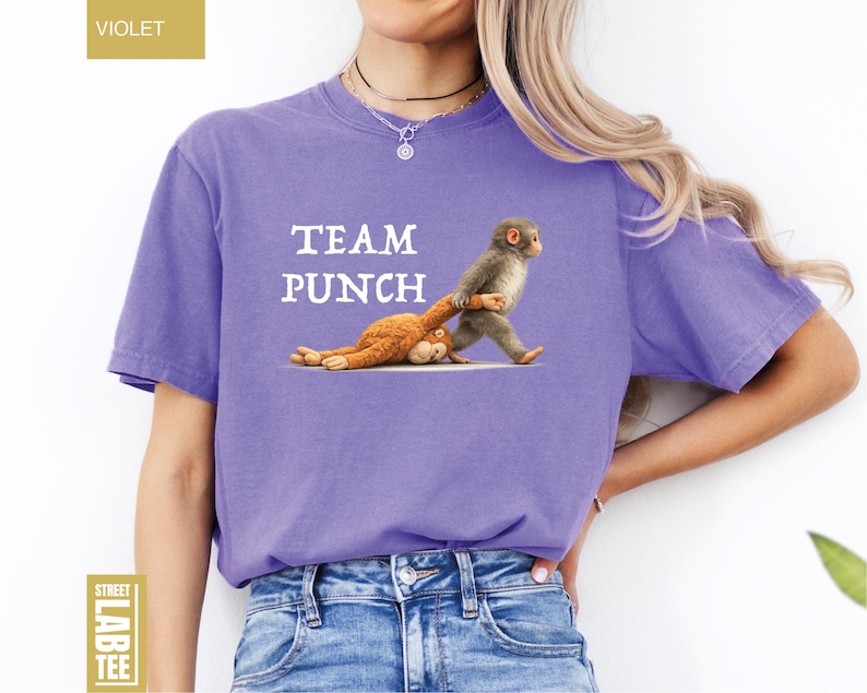 May include: A violet-colored t-shirt with the words "TEAM PUNCH" printed in white. The shirt features a graphic of a monkey pulling a stuffed animal. The shirt is made of a soft material.