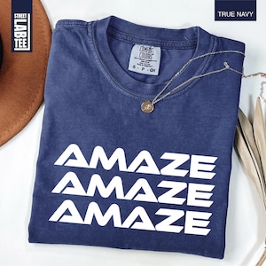 May include: A navy blue t-shirt with the word "AMAZE" printed three times in white. The shirt has a crew neck and short sleeves. A gold necklace with a pendant rests on the shirt. The shirt is labeled "TRUE NAVY".