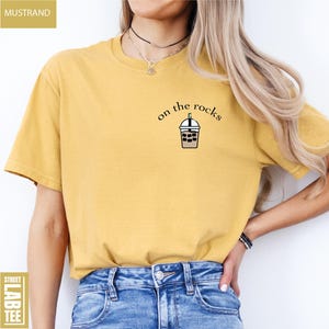 May include: Mustard-colored t-shirt with the text "on the rocks" and a graphic of an iced coffee. The shirt is a short-sleeved, crew neck style. The words "Street Lab Tee" are printed on a label.