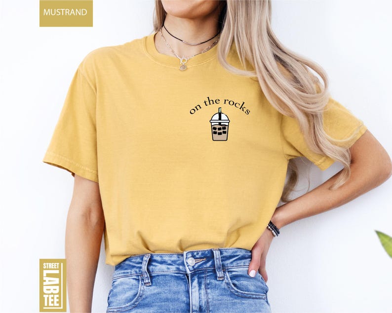 May include: Mustard-colored t-shirt with the text "on the rocks" and a graphic of an iced coffee. The shirt is a short-sleeved, crew neck style. The words "Street Lab Tee" are printed on a label.