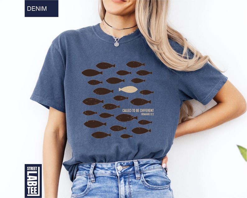 Puede incluir: Camiseta azul denim con un gr&aacute;fico de peces. El dise&ntilde;o presenta varios peces marrones nadando en una direcci&oacute;n, con un pez beige nadando en la direcci&oacute;n opuesta. El texto en la camiseta dice "CALLED TO BE DIFFERENT ROMANS 12:2".