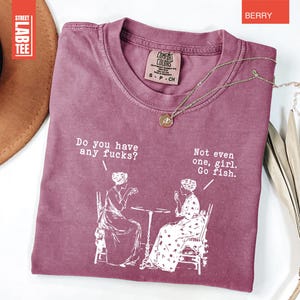 May include: Berry-colored t-shirt with the text "Do you have any fucks?" and "Not even one, girl. Go fish." in white. The graphic depicts two women seated at a table. A gold necklace is draped across the shirt.