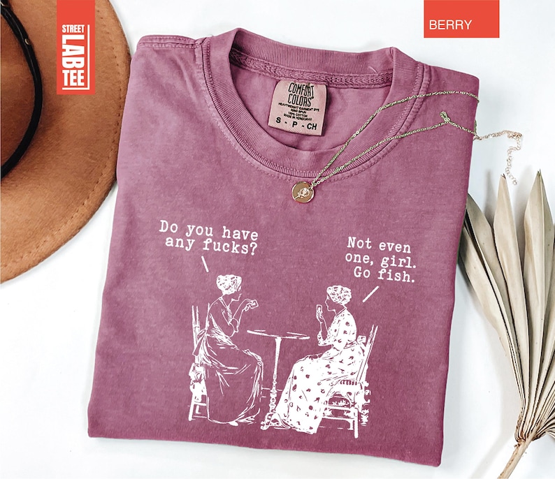May include: Berry-colored t-shirt with the text "Do you have any fucks?" and "Not even one, girl. Go fish." in white. The graphic depicts two women seated at a table. A gold necklace is draped across the shirt.