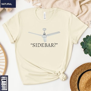 May include: Cream-colored t-shirt featuring a ceiling fan graphic and the text "SIDEBAR?". The shirt is made of a soft material and has a classic crew neck design. A casual and comfortable tee.