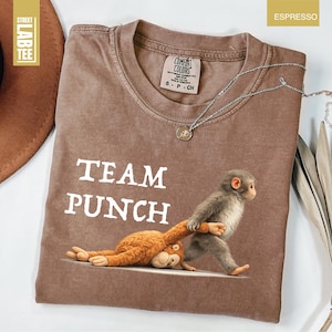 May include: Espresso-colored t-shirt with the text "TEAM PUNCH" and a graphic of a monkey pulling a stuffed animal. The shirt has a crew neck and a small tag near the collar. A silver necklace with a pendant is draped across the shirt.
