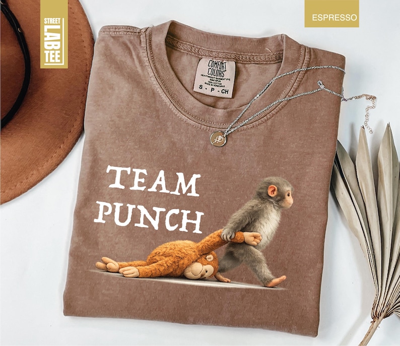 May include: Espresso-colored t-shirt with the text "TEAM PUNCH" and a graphic of a monkey pulling a stuffed animal. The shirt has a crew neck and a small tag near the collar. A silver necklace with a pendant is draped across the shirt.