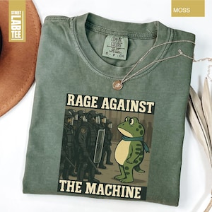 May include: Moss green t-shirt with the text "RAGE AGAINST THE MACHINE" and a graphic of a frog facing a line of riot police. The shirt has a crew neck and short sleeves. A gold necklace is draped on the shirt.