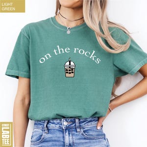 May include: Light green t-shirt featuring the words "on the rocks" and an iced coffee graphic. The shirt has short sleeves and a solid colour. The label reads "Street Lab Tee".