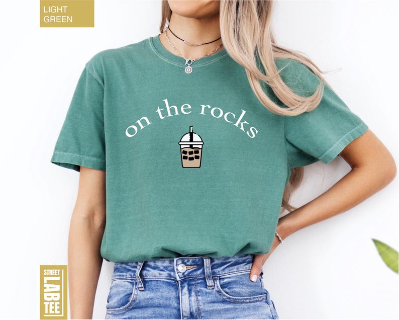May include: Light green t-shirt featuring the words "on the rocks" and an iced coffee graphic. The shirt has short sleeves and a solid colour. The label reads "Street Lab Tee".