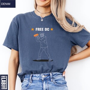Op de afbeelding: Denim blauw T-shirt met de tekst "FREE DC" en een afbeelding van een American footballspeler die een bal gooit. Het shirt heeft korte mouwen en een ronde hals.