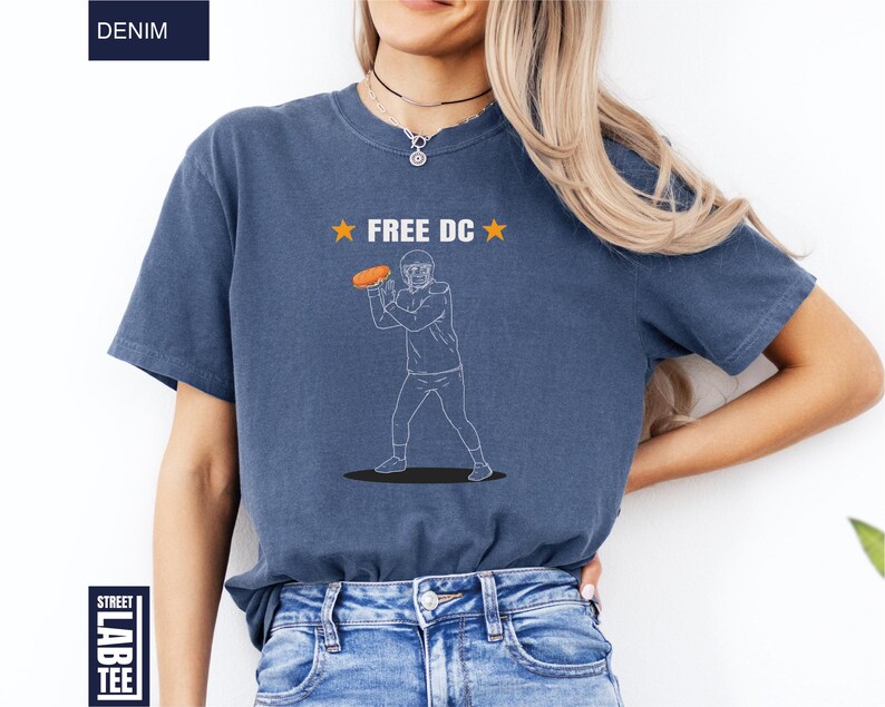 Op de afbeelding: Denim blauw T-shirt met de tekst "FREE DC" en een afbeelding van een American footballspeler die een bal gooit. Het shirt heeft korte mouwen en een ronde hals.