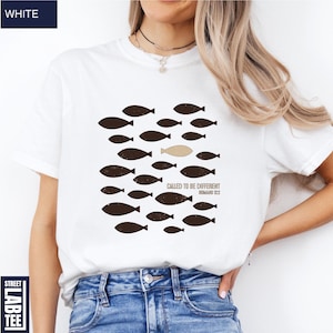 Puede incluir: Camiseta blanca con un gr&aacute;fico de peces marrones, uno beige, nadando juntos. El texto "CALLED TO BE DIFFERENT ROMANS 12:2" est&aacute; impreso debajo. La camiseta est&aacute; etiquetada como "WHITE" en la esquina superior izquierda.